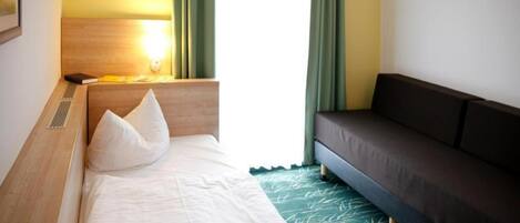 Une chambre d’hôtel comprenant un lit, un canapé, une tête de lit, une table de chevet avec une lampe et une fenêtre avec des rideaux.