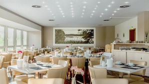 A modern dining area with round tables, beige chairs, and a large wall-mounted screen displaying a nature scene. - Kaiserhof Feldberg by Belvilla Resorts (Feldberger Seenlandschaft)