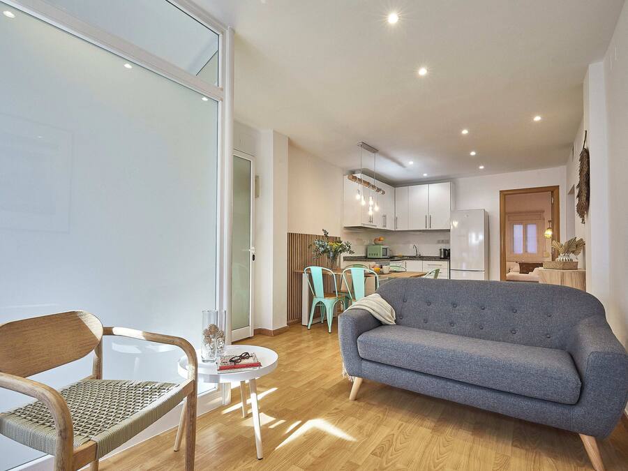 Una sala de estar moderna con un sofá gris, sillas de madera, una mesita redonda y una cocina al fondo.