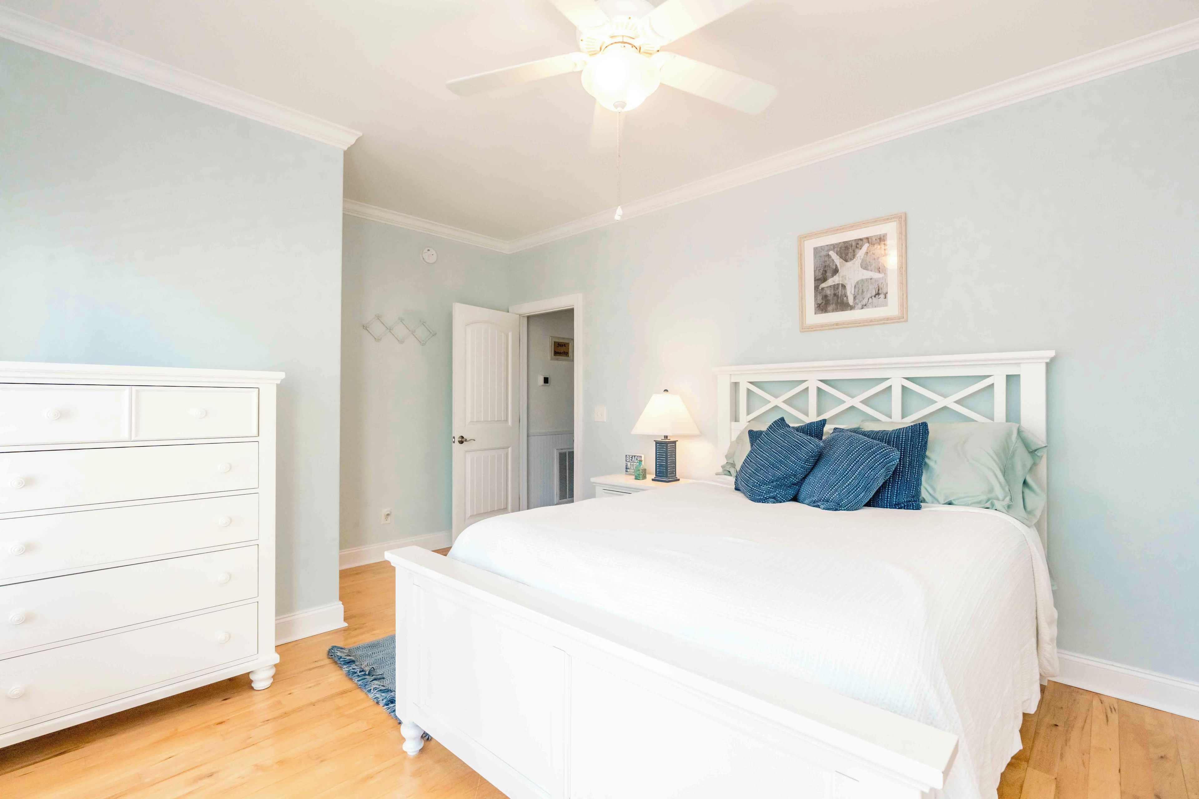A bedroom with a white bed, a dresser, a ceiling fan, and a door leading to another room.