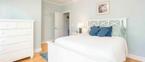 A bedroom with a white bed, a dresser, a ceiling fan, and a door leading to another room.