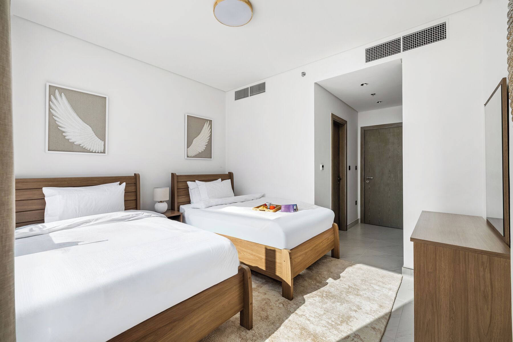 Ein Hotelzimmer mit zwei Betten, einem hölzernen Kopfteil, einem Nachttisch mit Lampe und einem kleinen Tablett mit Frühstücksangeboten.