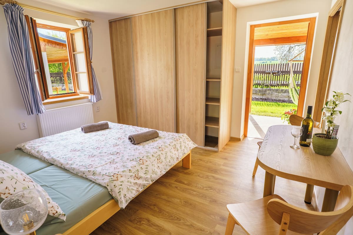 A bedroom with a bed, a wooden table with a chair, a window with curtains, and a view of the outdoors.