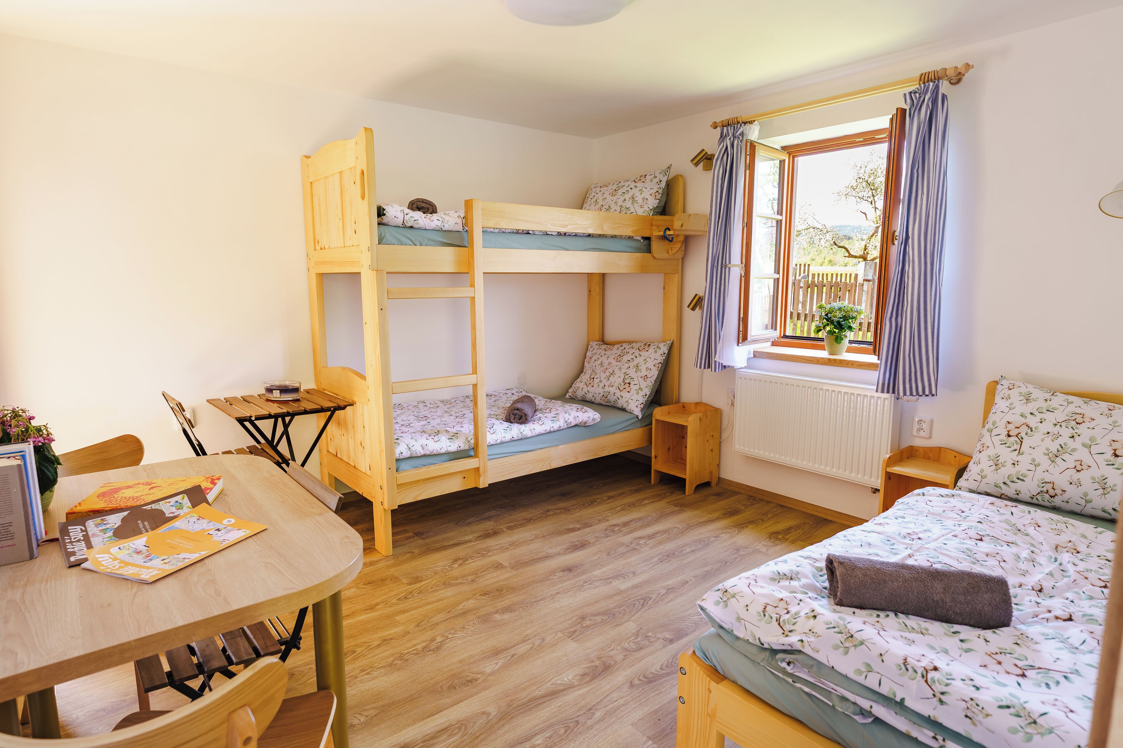 Una habitación con literas, una mesa de madera, una ventana con cortinas y un radiador.