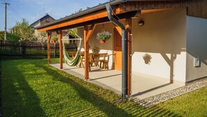 A covered patio area with a wooden table and chairs, a hammock, and hanging flower baskets. - Vlaštovčí recreation area (Radvanec)