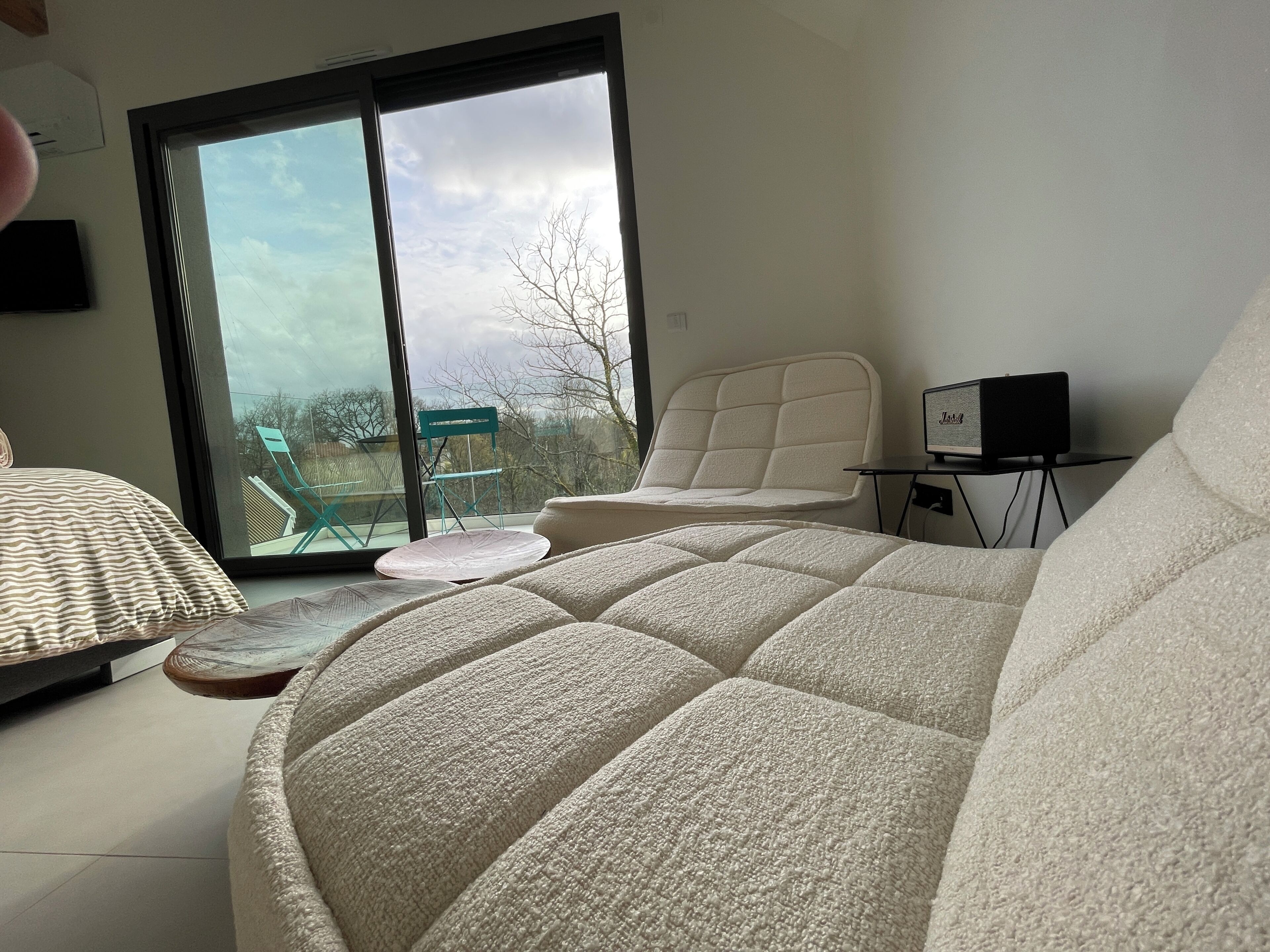 Ein modernes Schlafzimmer mit einem großen Bett, einem Sessel, einem kleinen Tisch mit einem Lautsprecher und Blick durch eine Schiebetür.