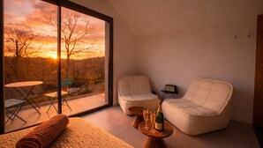 Een moderne kamer met een groot raam, twee comfortabele fauteuils, een tafeltje met een fles en glazen, en uitzicht op een zonsondergang.