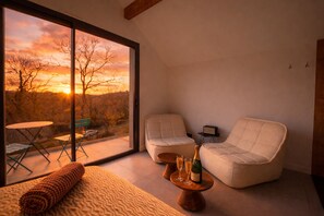 A modern room with a large window, two cushioned chairs, a small table with a bottle and glasses, and a view of a sunset.