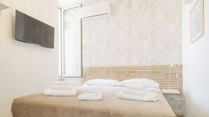 Un lit bien fait, recouvert d’une couverture beige, un téléviseur à écran plat fixé au mur et une fenêtre avec des stores.