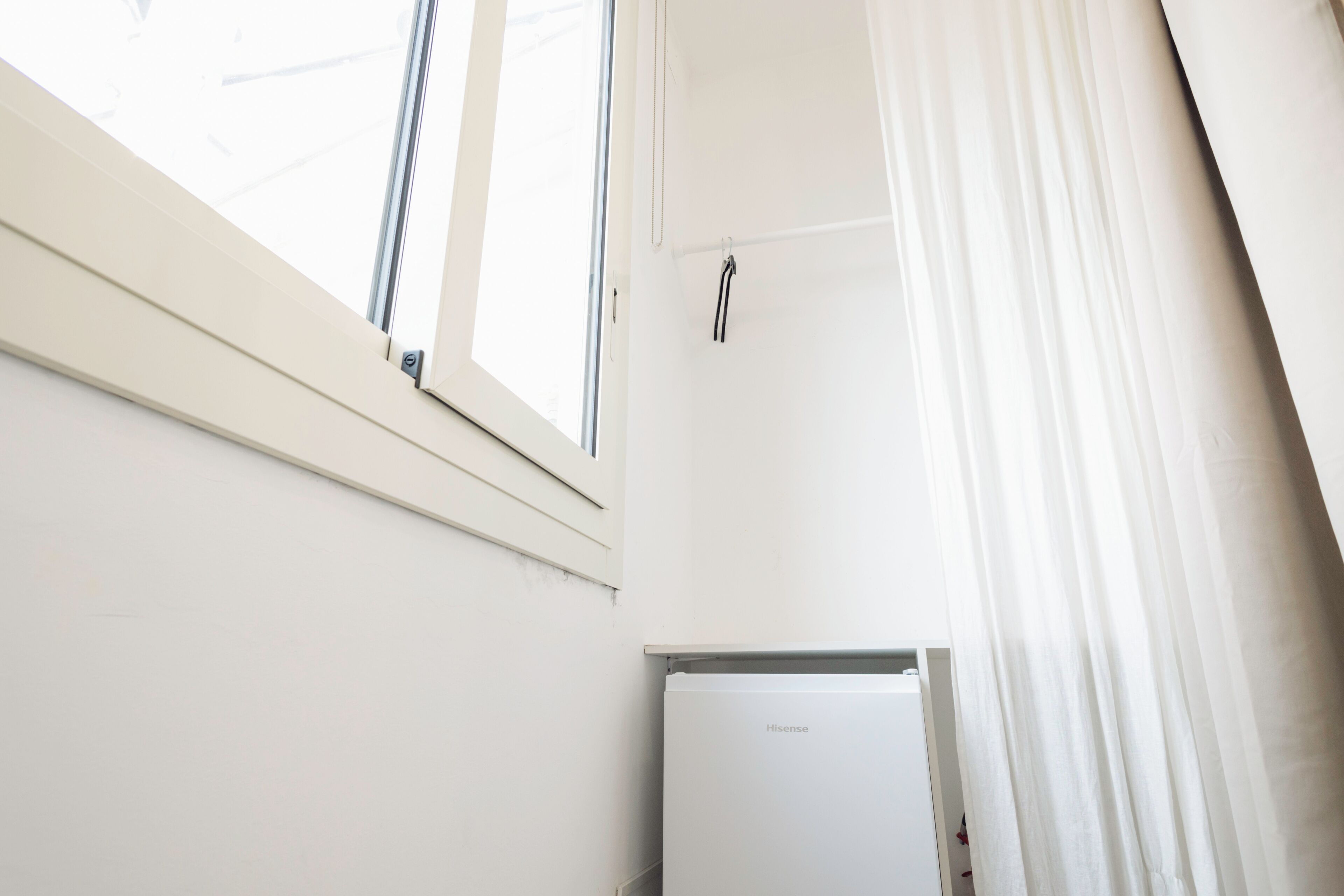 Ein Zimmer mit weißer Wand, einem Fenster mit Jalousien und einem weißen Kühlschrank.