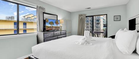 A hotel room with a large bed, a TV, a dresser, and a balcony with a view of other buildings.