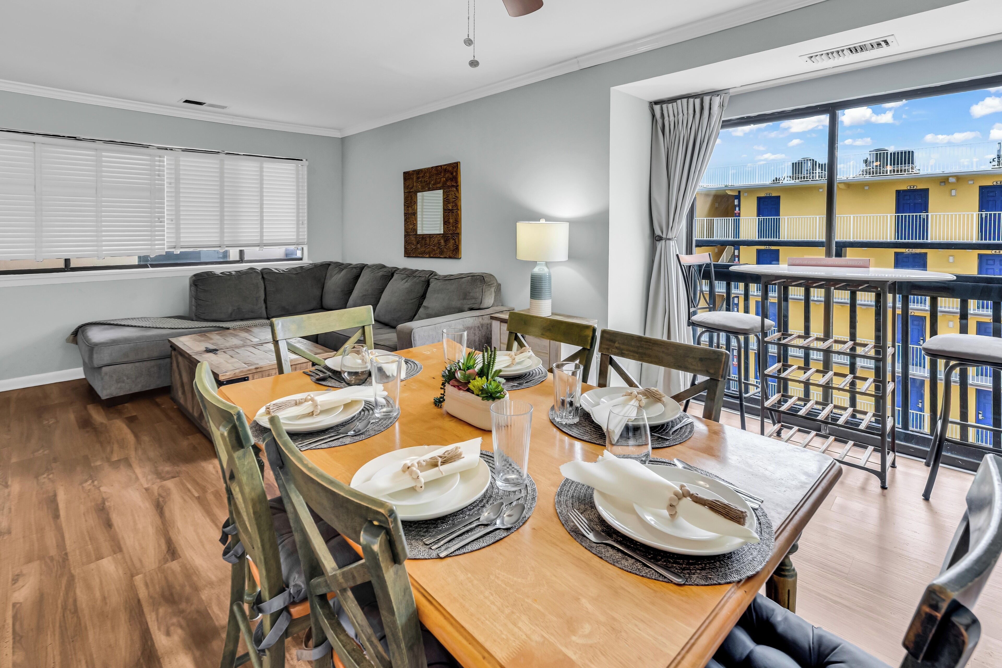 Un coin repas avec une table en bois dressée pour quatre personnes, un canapé gris et un balcon aux murs jaunes et aux balustrades bleues.