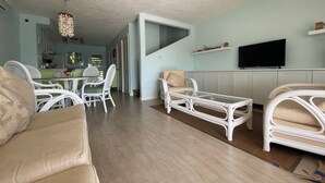 Un salón espacioso con techo alto, muebles de mimbre blanco, un televisor de pantalla plana montado en la pared y una zona de comedor al fondo.