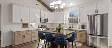 A modern kitchen with white cabinets, a stainless steel refrigerator, a central island with bar stools, and a chandelier.