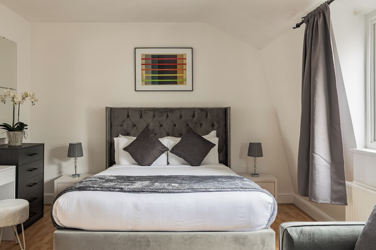 Camera da letto con testiera trapuntata, due comodini con lampade, un letto con coperta a righe bianche e grigie, un quadro astratto incorniciato sopra il letto e una finestra con tende.