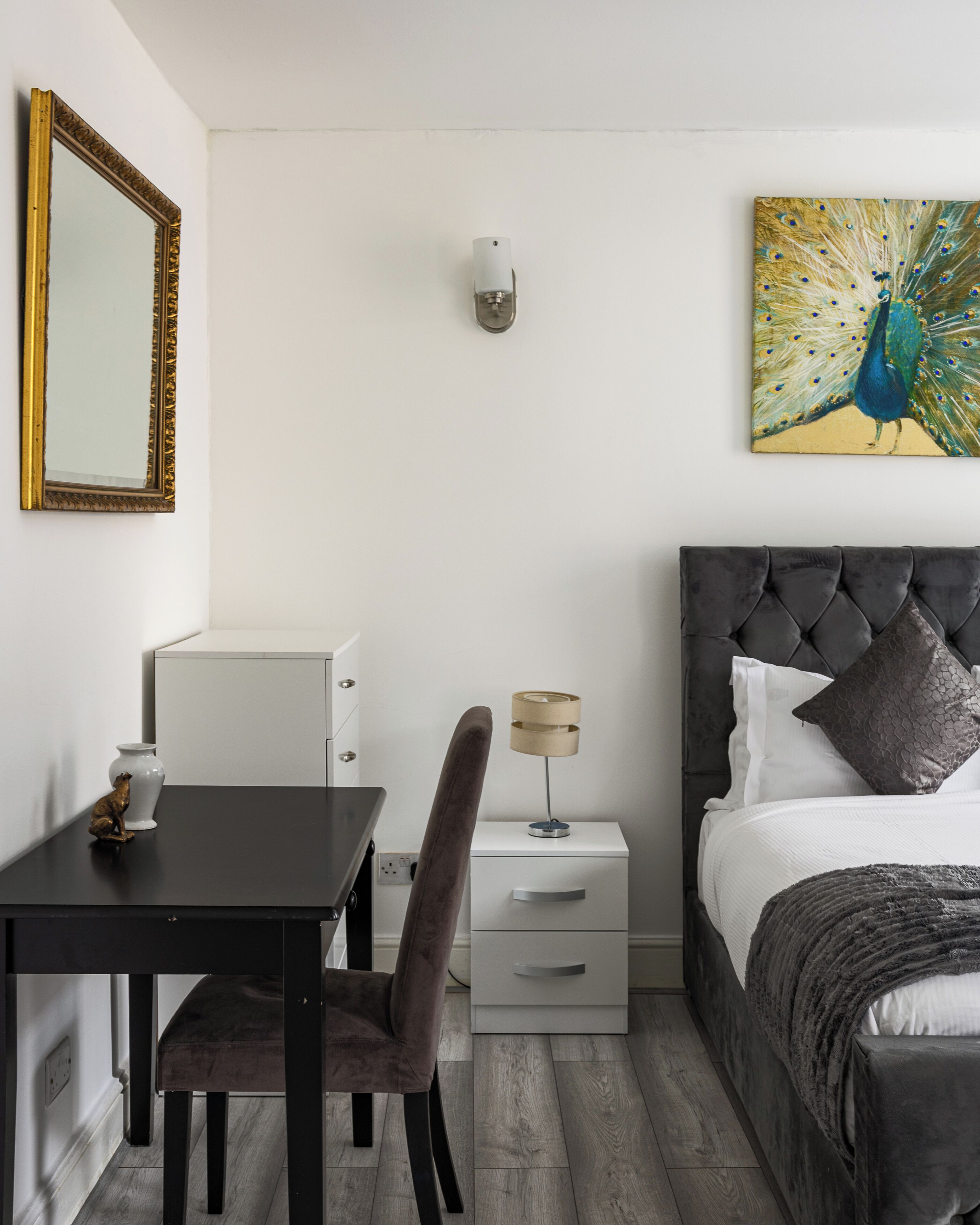 A hotel room with a bed, a desk with a chair, a nightstand with a lamp, a wall-mounted mirror, and a peacock painting.