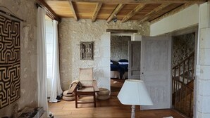 Una habitación rústica con vigas de madera a la vista, una pared de piedra, una silla de mimbre, una mesa de madera y una lámpara blanca.