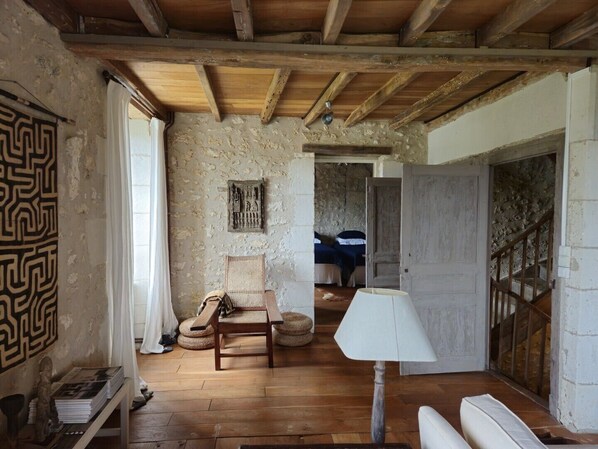 A rustic room with exposed wooden beams, a stone wall, a woven chair, a wooden table, and a white lamp.