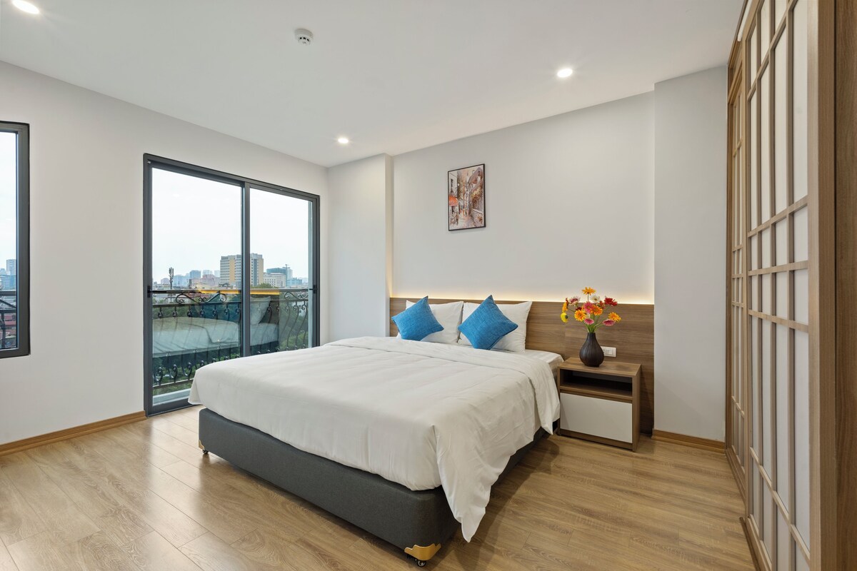 A modern bedroom with a large bed, a nightstand, a vase with flowers, and a city view through a sliding door.