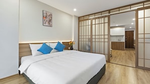 A modern hotel room with a large bed, a wooden headboard, a nightstand with a vase of flowers, and a sliding door leading to a kitchen area. - Residence Suite (Hanoi)