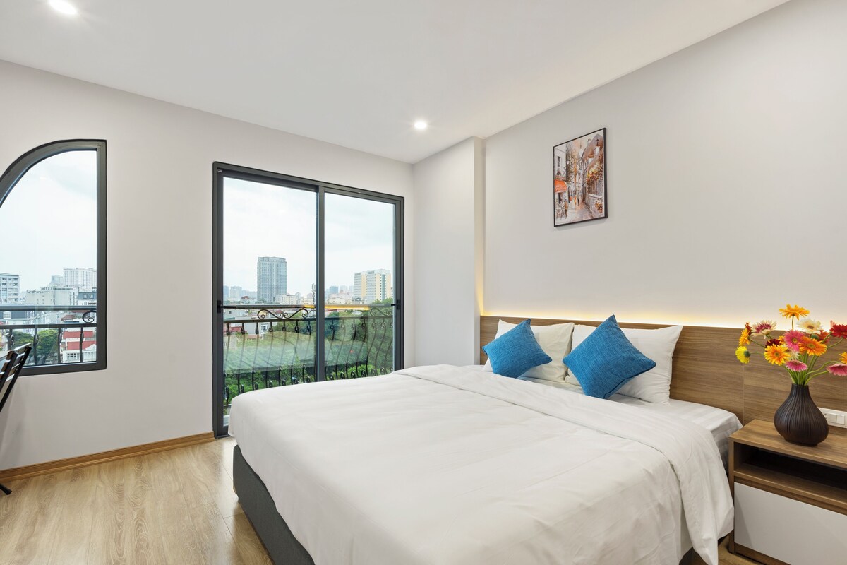 A modern hotel room with a large bed, a city view through a balcony, and a small table with a vase of flowers.