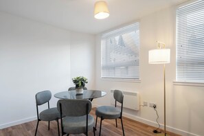 A modern dining area with a round glass table, four grey chairs, a small potted plant, and a floor lamp. - City Point Apartments by Guestz (Aberdeen)