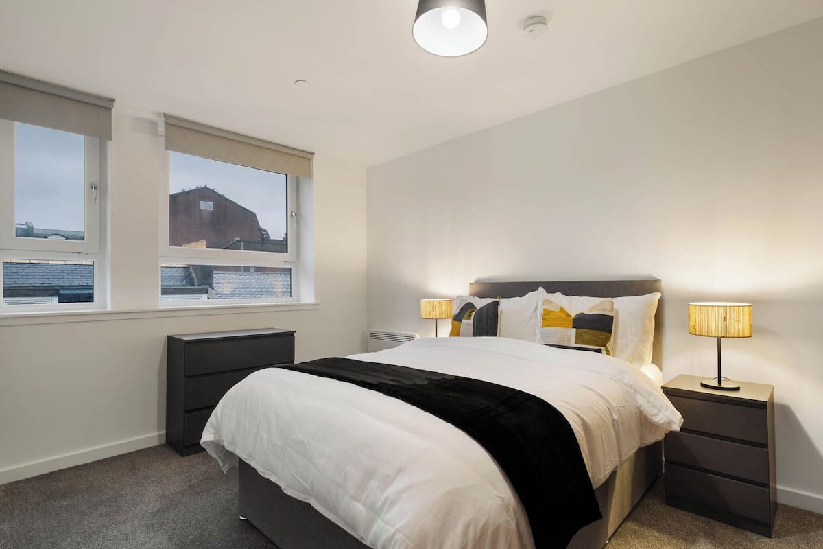 Un dormitorio con cama, dos mesitas de noche, una ventana y vista a un edificio.