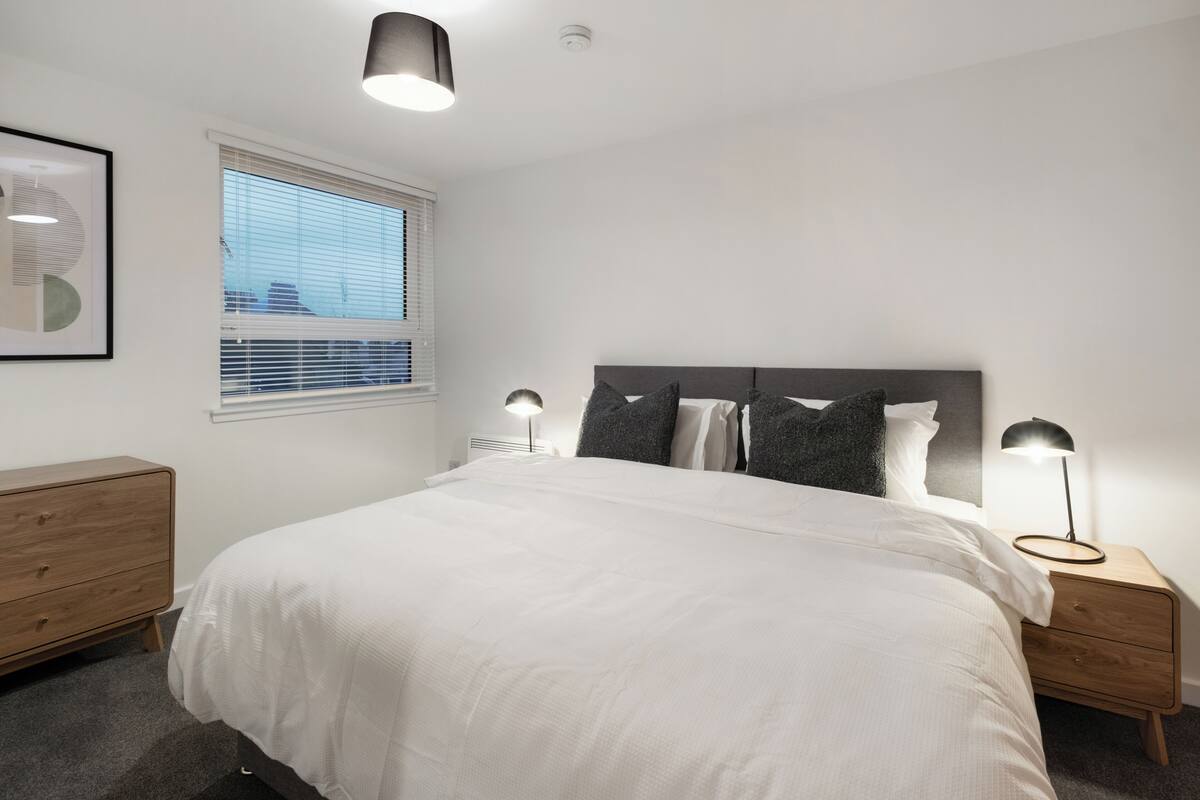 Un dormitorio con cama, lámparas de noche, mesita de noche, cómoda, ventana con persianas y lámpara de techo.