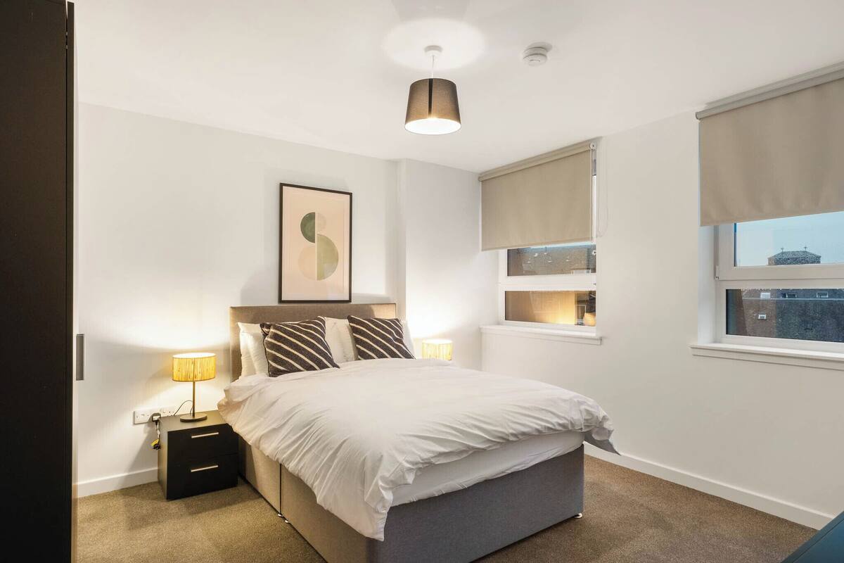 Un dormitorio con cama, mesitas de noche, lámpara, cuadro enmarco, ventana con persianas y luz de techo.