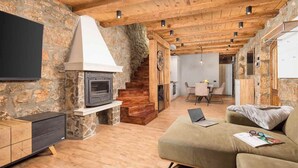 A cozy living room with a stone wall, wooden ceiling, and a fireplace. There is a sofa, a TV, and a dining area in the background. - Private Stone Villa with heated pool, Near Opatija (Ičići)