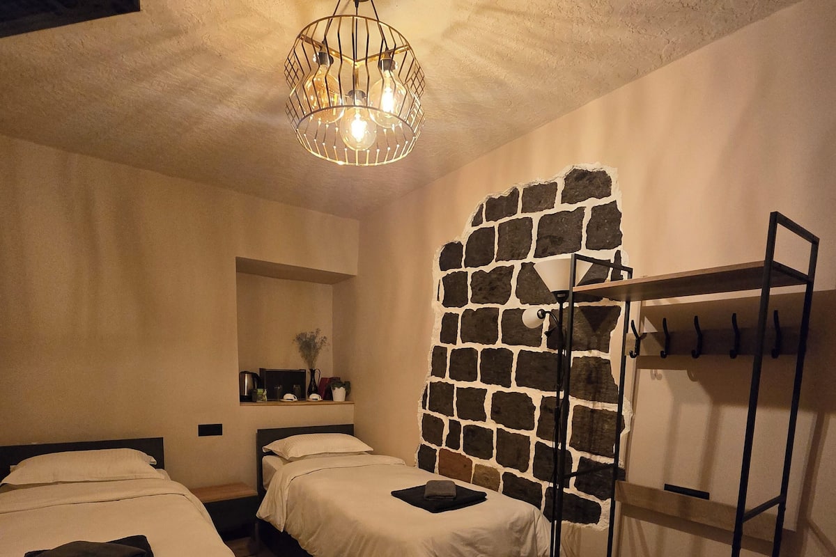 A compact hotel room with two beds, a wooden table, a chair, a hanging light fixture, and a stone accent wall.