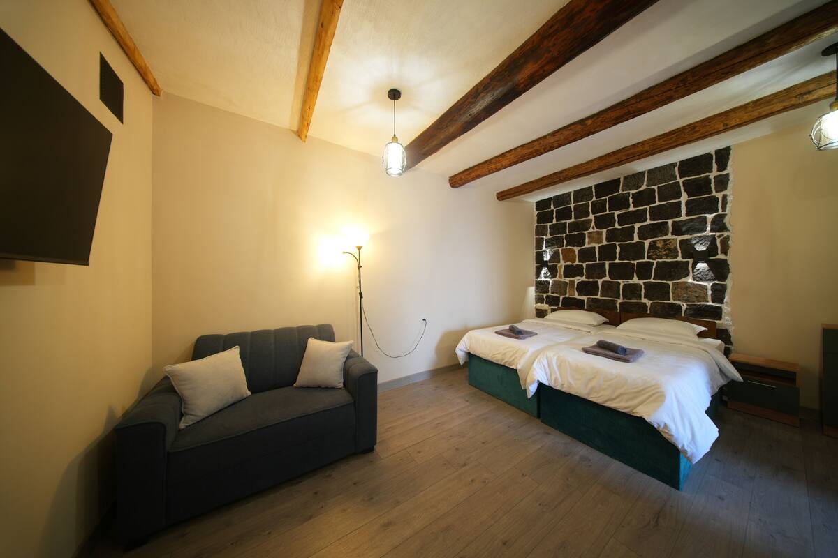 A bedroom with a stone headboard, a bed with white bedding, a dark sofa with cushions, and a wall-mounted light fixture.