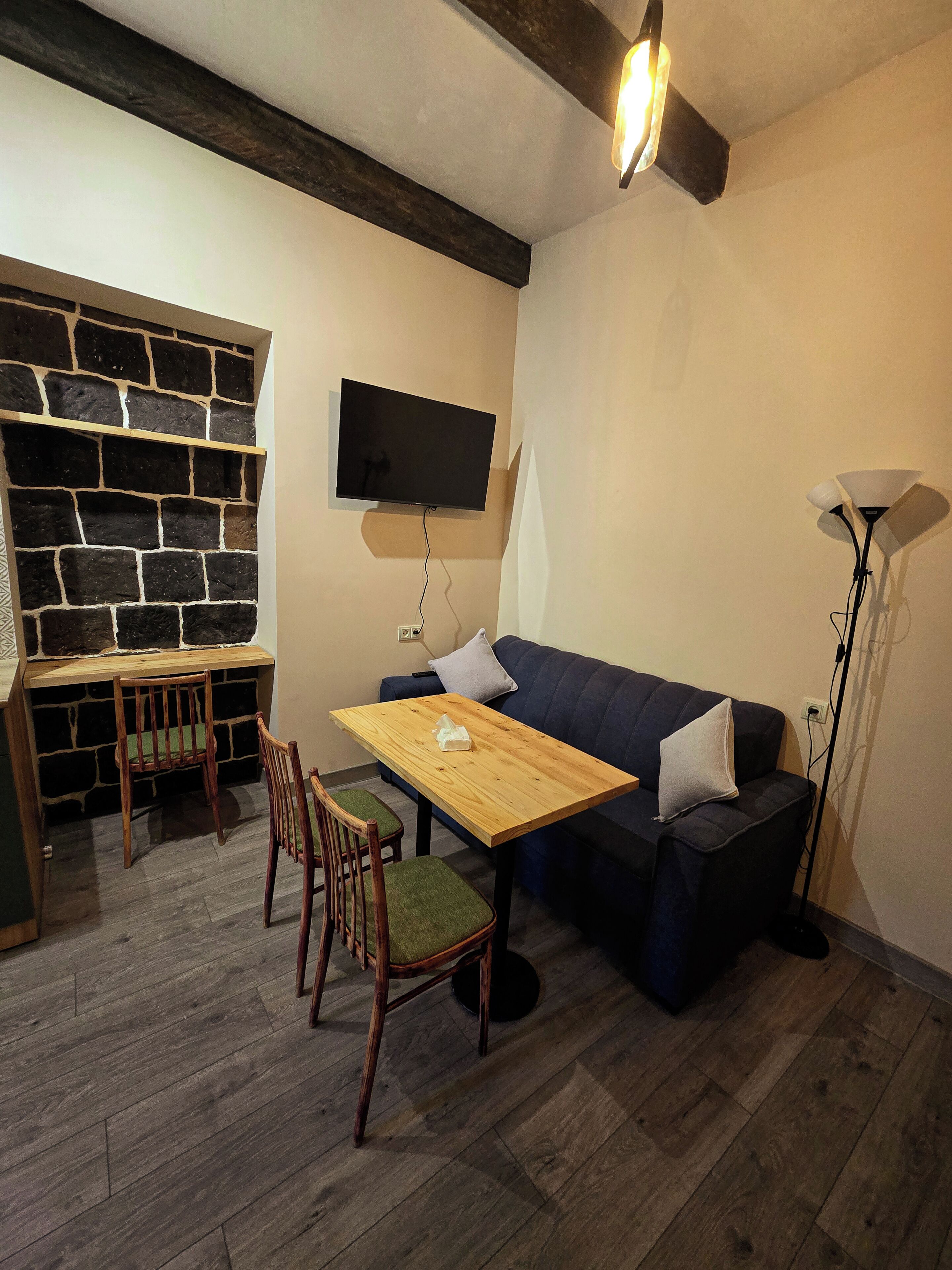 A cozy living room with a stone wall, a flat-screen TV, a wooden table with chairs, a dark blue sofa, and a floor lamp.