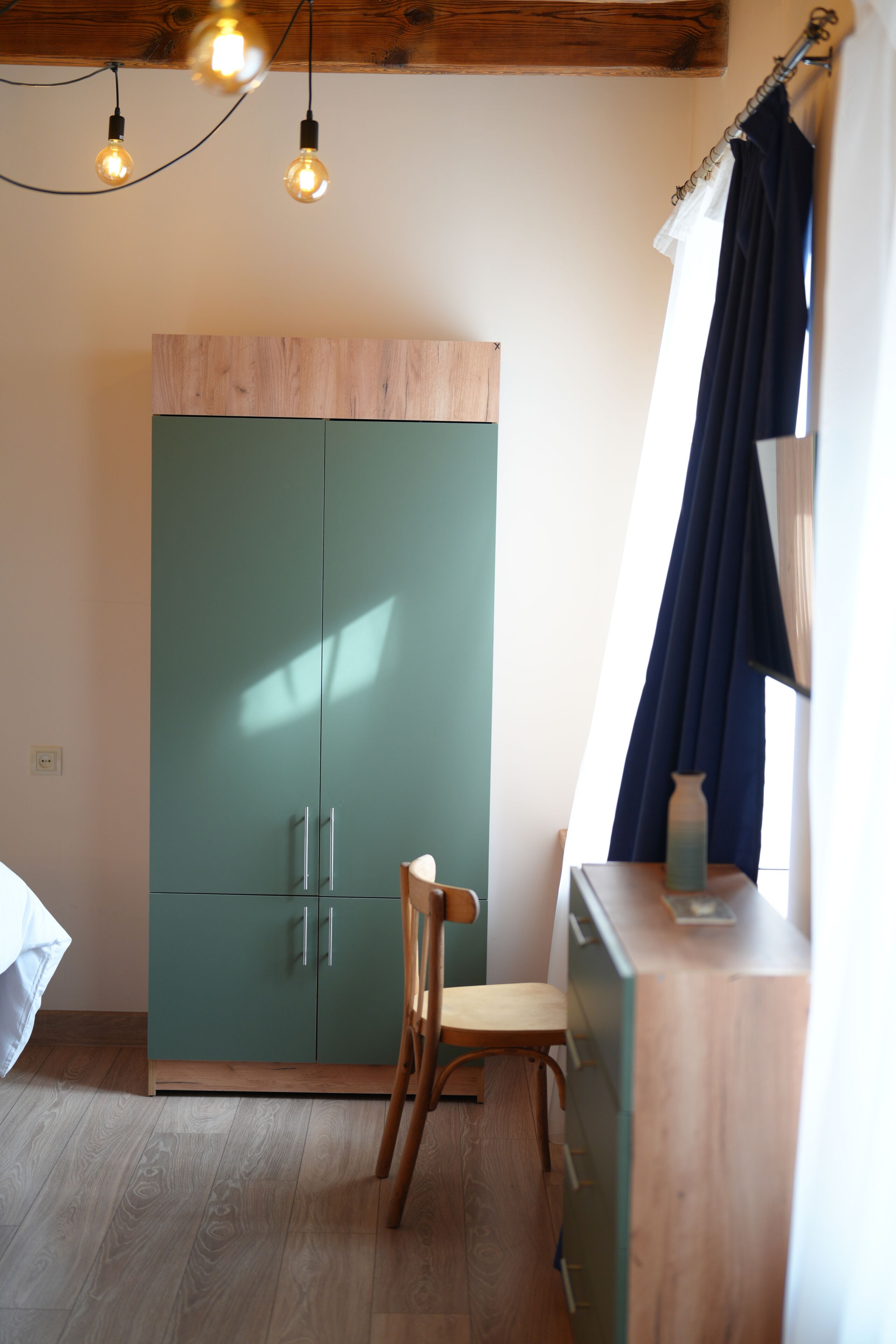 A room with a wooden ceiling, a green wardrobe, a wooden chair, and a window with curtains.