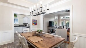 Un coin repas avec une table en bois, des chaises blanches, un lustre et une vue sur la cuisine et le salon.