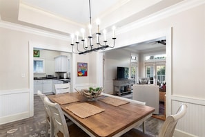 Un coin repas avec une table en bois, des chaises blanches, un lustre et une vue sur la cuisine et le salon.