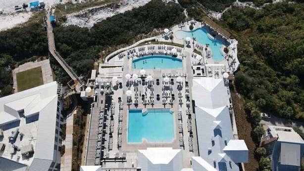 Vue aérienne d’un complexe hôtelier doté de plusieurs piscines, d’un parking et d’un grand bâtiment dont le toit présente un design géométrique original.