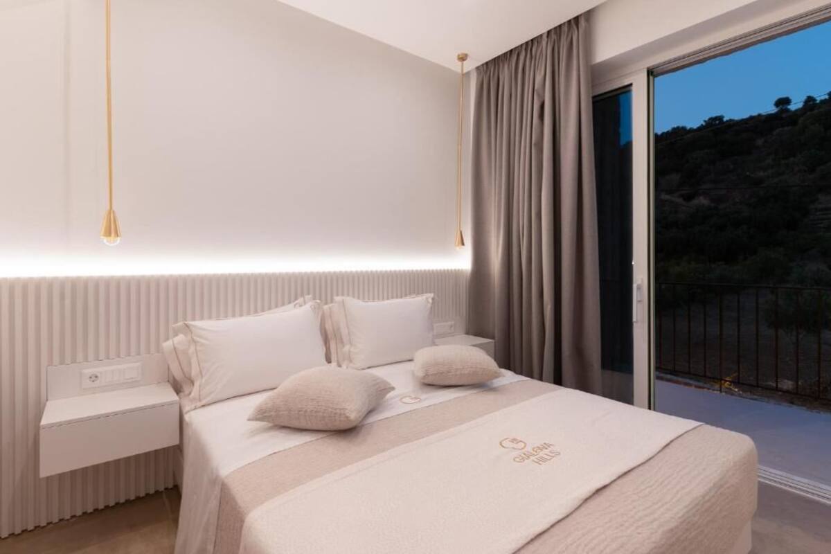 A modern hotel room with a large bed, a nightstand, and a view of the outdoors.
