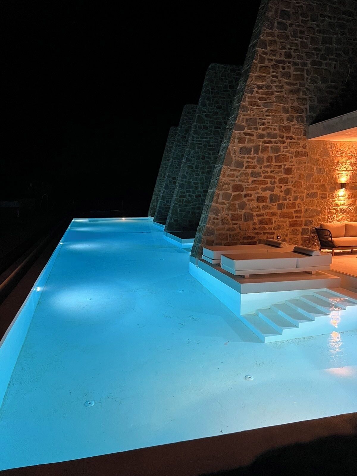 A swimming pool with stone walls and a seating area with cushions.