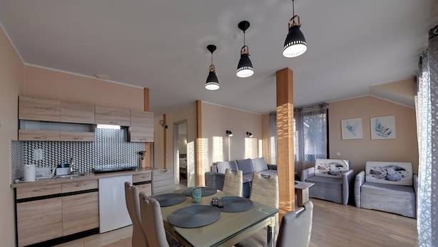 A modern kitchen with wooden cabinets, a dining area with a glass table, and a living room with sofas and artwork. - RentPlanet - Apartamenty przy Wyciągu (Szklarska Poręba)