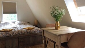 Un dormitorio con techo inclinado, una cama con ropa de cama estampada, una mesa de madera con un jarrón de flores y una ventana con persianas.