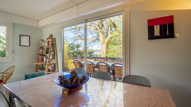 Un comedor con una mesa grande, sillas, un cuenco de piñas, una estantería y vistas al exterior.
