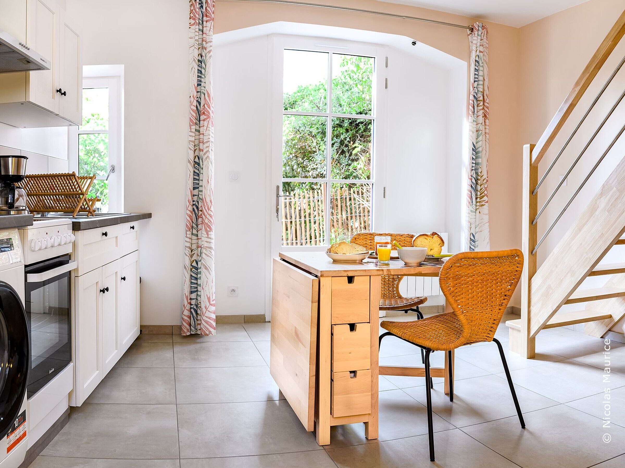 A compact kitchen with white cabinets, a built-in oven, and a coffee machine. There's a dining area with a wooden table, a wicker chair, and a window with a view of greenery.