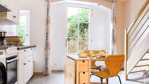 Une cuisine compacte avec des meubles blancs, un four encastré et une machine à café. On y trouve un coin repas avec une table en bois, une chaise en osier et une fenêtre donnant sur un paysage verdoyant.