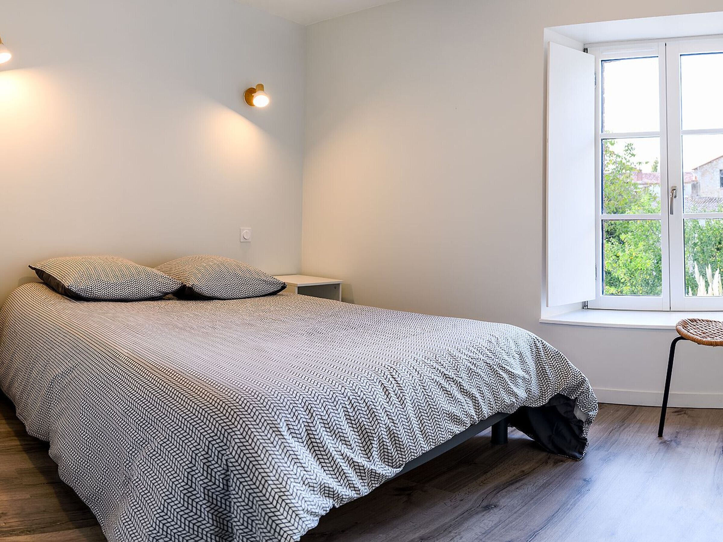 A bedroom with a bed, two pillows, a nightstand, a chair, and a window with a view of greenery.