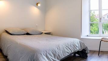 A bedroom with a bed, two pillows, a nightstand, a chair, and a window with a view of greenery.