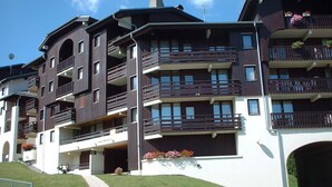 A multi-story building with balconies, a paved driveway, and a sign indicating access to a nearby golf course. - Lovely 1-bedroom apartment, mountain corner, balcony, pets allowed (Morillon Village)