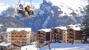 A ski lift with skiers, a mountain backdrop, and a ski resort building with balconies. - Lovely 1-bedroom apartment, mountain corner, balcony, pets allowed (Morillon Village)