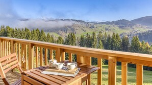 Eine Holzterrasse mit einem für Tee gedeckten Tisch, mit Blick auf eine gebirgige Landschaft mit einem bewaldeten Tal und einem See.