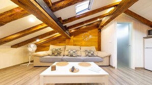 Un dormitorio estilo loft acogedor, con techo de madera, un sofá con cojines estampados, una mesita de centro blanca y un pequeño ventilador.